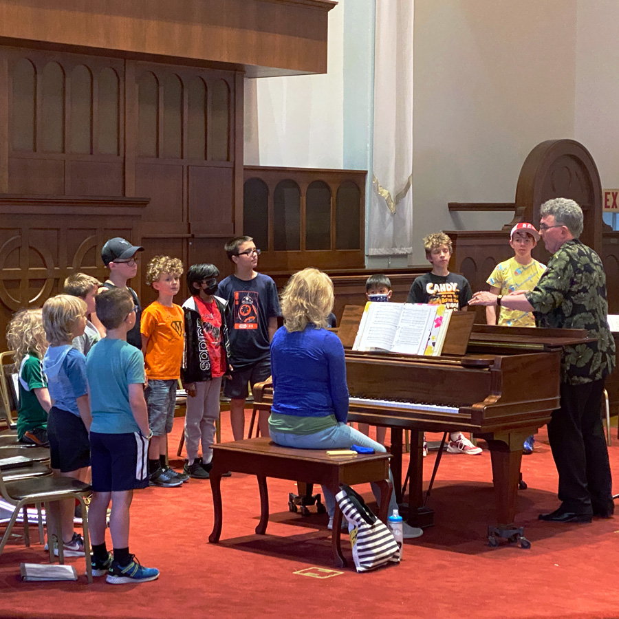 La Crosse Boychoir kids rehearsing a song around a piano and conductor at Cappella Performing Arts Center in Wisconsin.