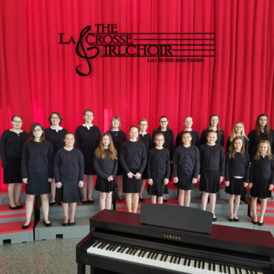 La Crosse Girlchoir dressed in formal black on stage in front of a red curtain.